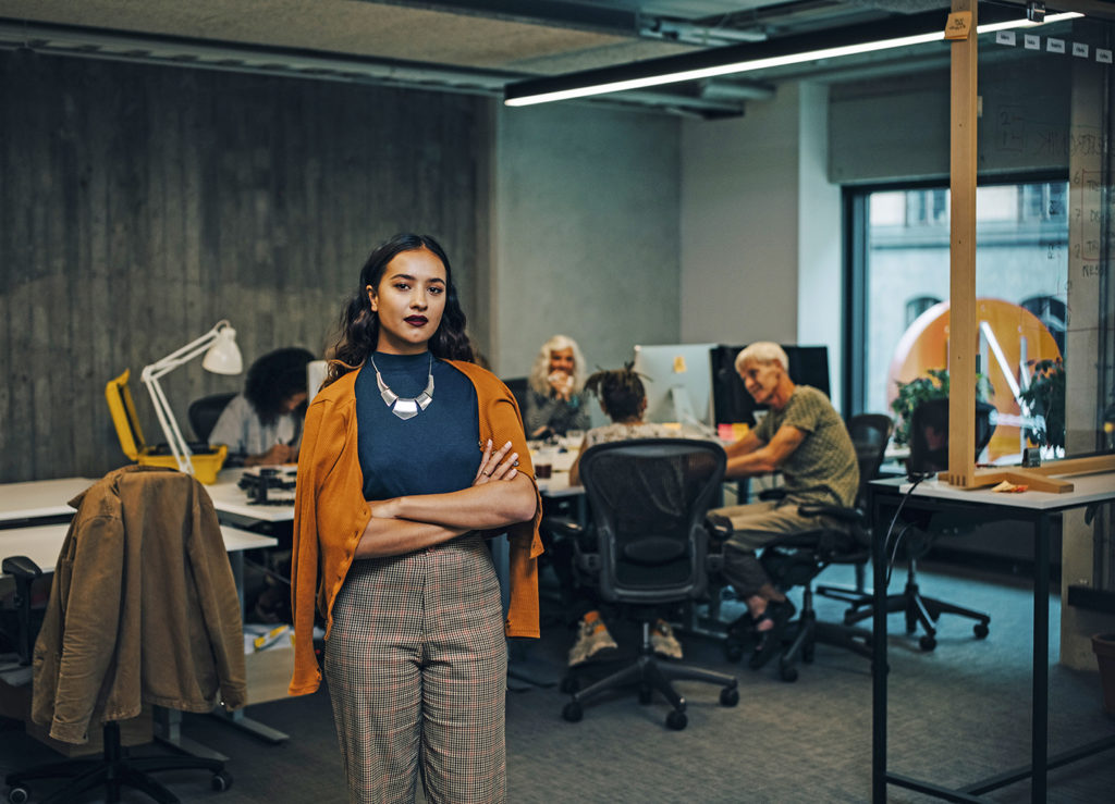 Confident businesswoman with arms crossed while colleagues sitting in background at office